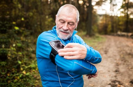 Senior runner in nature. Man with smartphone and earphones, adjusting settings on armband for phone. Listening music or using a fitness app. Using phone app for tracking weight loss progress, running goal or summary of his run.